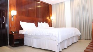 A hotel room with a large bed, wooden headboard, bedside lamps, a nightstand, and a wardrobe. - Sainte Famille Hotel (Kigali)