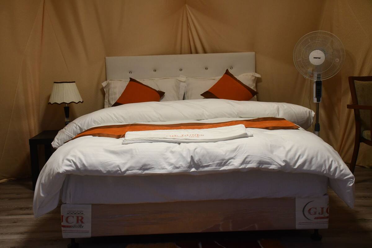 Ein ordentlich bezogenes Bett mit weißer und orangefarbener Bettwäsche, eine Nachttischlampe, ein Standventilator und ein hölzerner Stuhl.