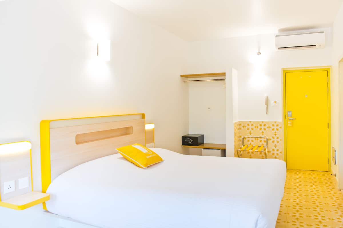 A hotel room with a bed, bedside table, lamp, and a yellow door.
