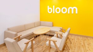A modern room with a beige sofa, a round wooden table, and four chairs. A bright yellow wall with the word "bloom" is visible. - Bloomrooms @ City Centre (Bengaluru)