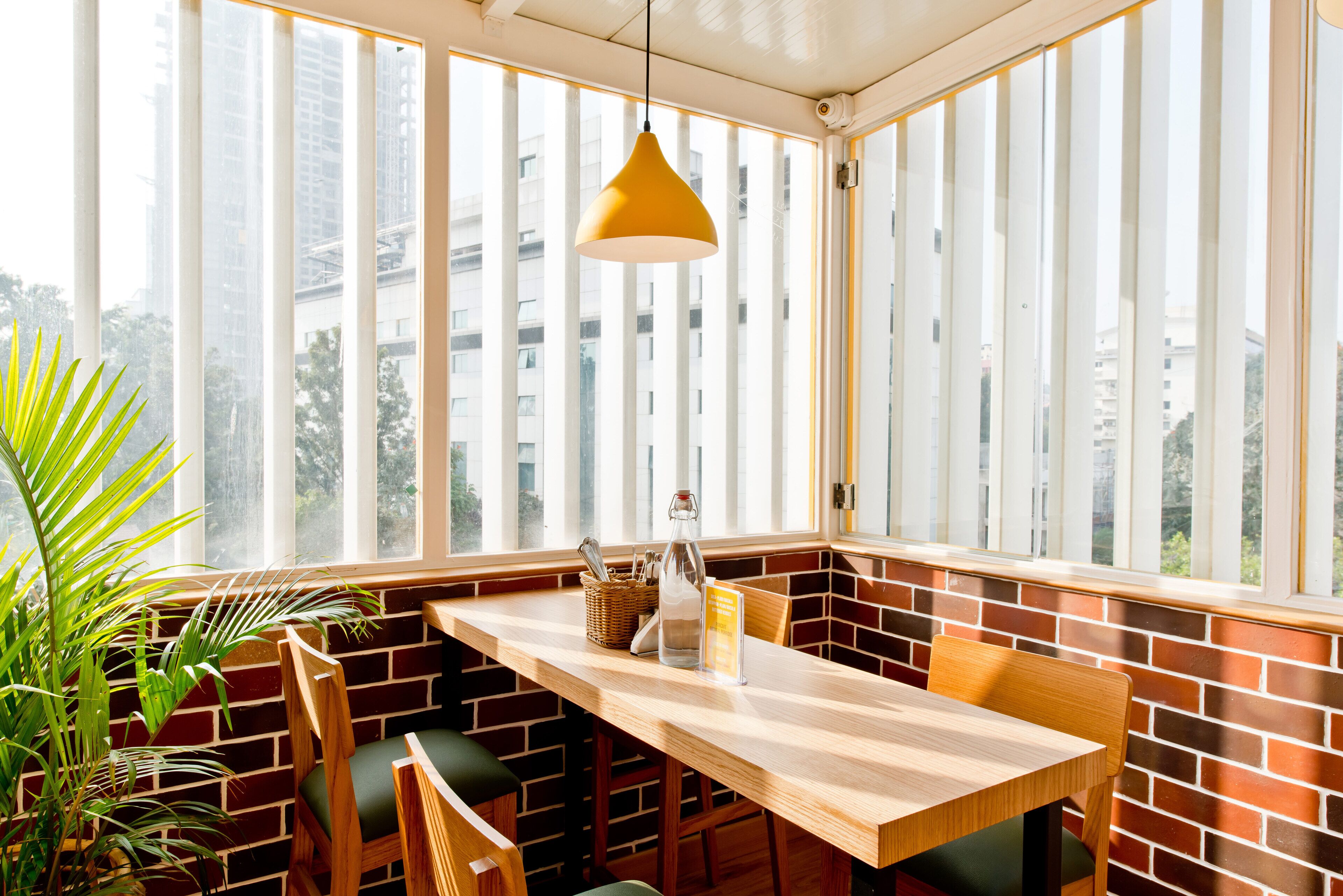 A corner seating area with a wooden table and chairs, a hanging pendant light, and a view of buildings through large windows.