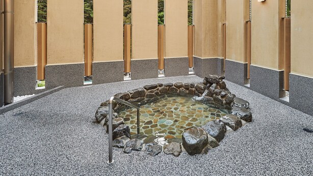 Un jardín interior tranquilo con una fuente de piedra, rodeado por un sendero de guijarros y un muro bajo con listones de madera verticales.