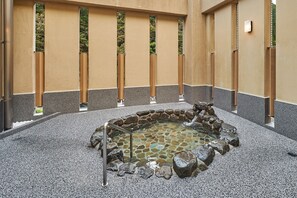 Un jardín interior tranquilo con una fuente de piedra, rodeado por un sendero de guijarros y un muro bajo con listones de madera verticales.