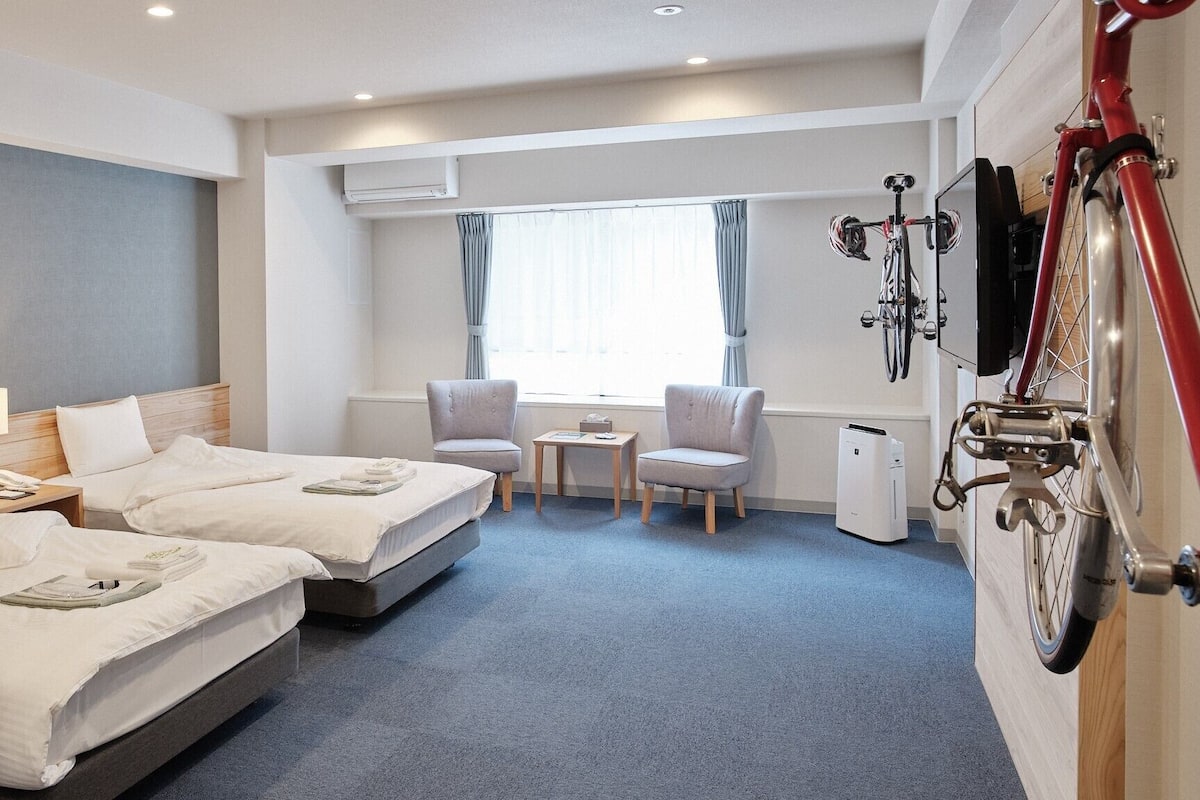 Habitación de hotel con dos camas, un escritorio, una silla, una bicicleta y una ventana con cortinas.