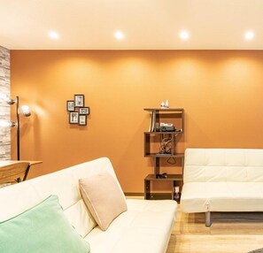 A modern living room with a beige sofa, a white sofa, and a small wooden side table. There is a wall-mounted shelf with framed pictures and a small shelf with electronic devices.