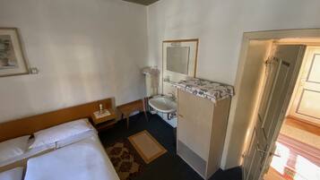 A hotel room with a bed, a bedside table, a small chair, a bathroom with a sink and mirror, and a door leading to another room.