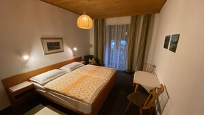 Quarto de hotel com teto de madeira, uma cama grande com colcha florida, uma cadeira, uma mesinha e uma janela com cortinas.