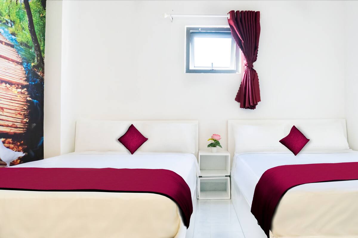 A hotel room with two beds, a small window with curtains, a nightstand with a vase of flowers, and a wall-mounted picture.