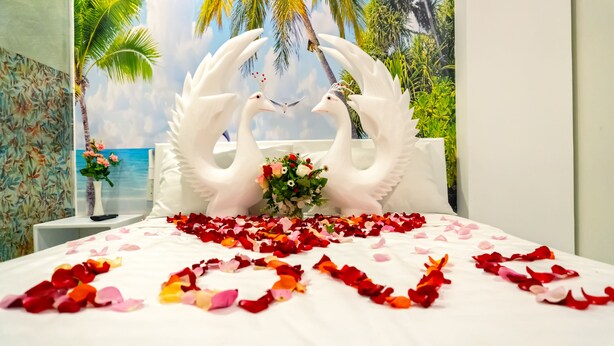 A bed with a white bedspread, two white swan-shaped pillows, a bouquet of flowers, and rose petals arranged on the bed. - Hoang Van Homestay Nha Trang (Bac Nha Trang)