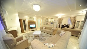 A living room with a sofa set, a television, a dining table with chairs, and a kitchen area in the background. - Helena Villa (Alanya)