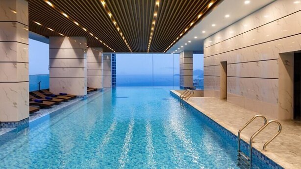 A modern indoor swimming pool with clear blue water, wooden slatted ceiling, and a view of the sea. - Apartment Beach Nha Trang Sea View (Nha Trang)