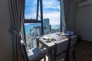 A dining area with a table set for two, large windows with curtains, and a view of a cityscape and water. - Apartment Beach Nha Trang Sea View (Nha Trang)