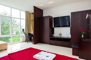 A modern hotel room with a large window, a bed with a red blanket, a TV mounted on the wall, and a desk with a chair.