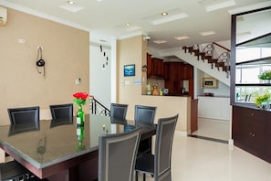 A dining area with a glass table and chairs, a kitchen in the background, and a staircase leading to another level.