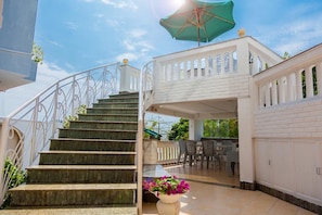 Una escalera conduce a un espacio exterior cubierto con una mesa y sillas, una valla blanca y un paraguas verde.