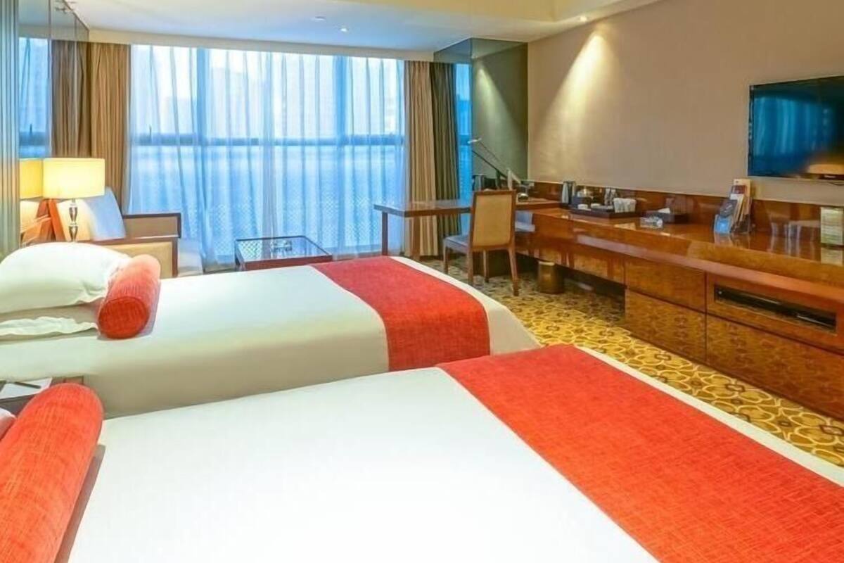 Hotelkamer met twee bedden, een bureau, een stoel, een televisie en een groot raam met gordijnen.