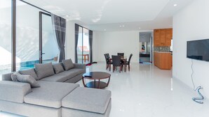 A modern living room with a large sectional sofa, a dining area with a table and chairs, and a kitchen area with wooden cabinets and a TV mounted on the wall. - Villa Viba (Ho Chi Minh City)