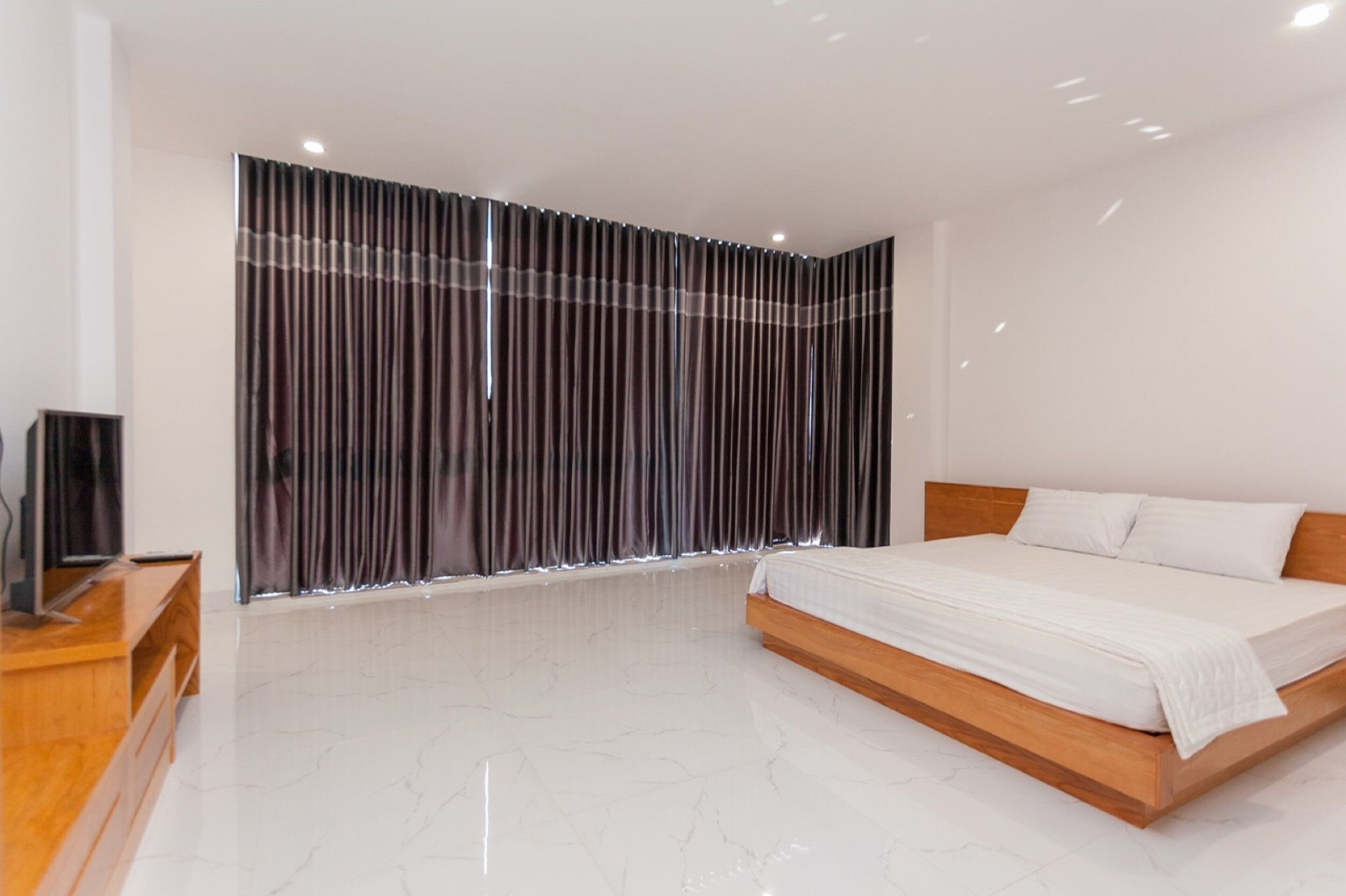 A modern bedroom with a wooden bed, a TV on a wooden stand, and a large window with vertical blinds.