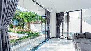 A modern living room with a large glass window, a grey sofa, and a view of a landscaped garden. - Villa Viba (Ho Chi Minh City)