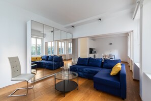 Un salon moderne avec un canapé bleu, une table basse en verre et un fauteuil blanc. Un grand miroir reflète la pièce, et on aperçoit un coin cuisine au fond.
