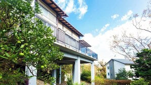 A two-story house with a balcony, surrounded by lush greenery and a clear sky.