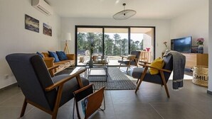 A modern living room with a sofa, armchairs, a coffee table, and a TV. There is a large window with a view of trees and a balcony. - Résidence Calarossa Bay Resort (Lecci)