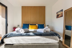 A neatly made bed with blue and yellow pillows, a grey blanket, and two rolled towels.