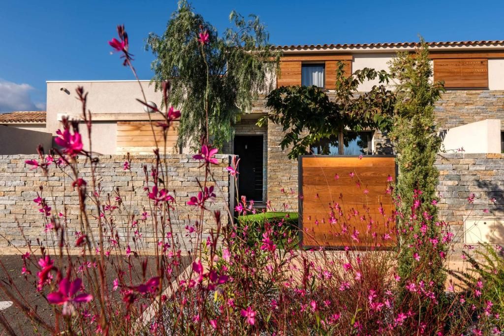 A modern house with a stone facade, a wooden door, and a small garden with pink flowers.