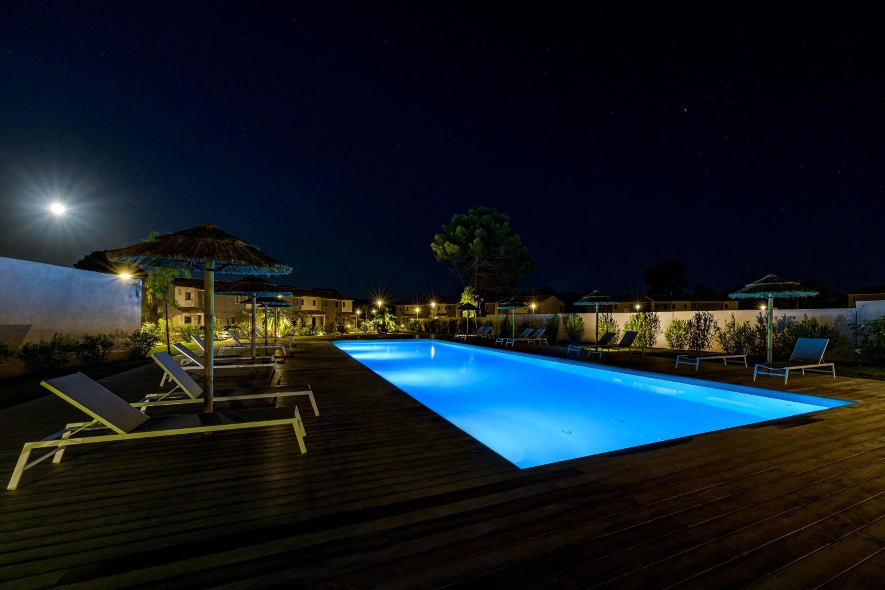 Une piscine nocturne, dotée de chaises longues et de parasols, entourée de bâtiments et sous un ciel dégagé.