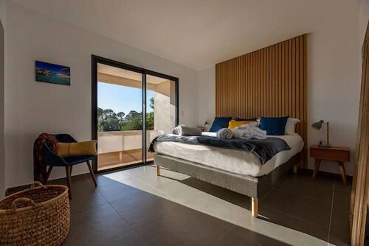 A modern bedroom with a large bed, a chair, a small table, and a view through a sliding glass door.