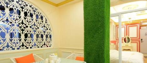 A room with a glass table surrounded by wicker chairs, a green artificial grass pillar, and a bed with a white and red patterned headboard.