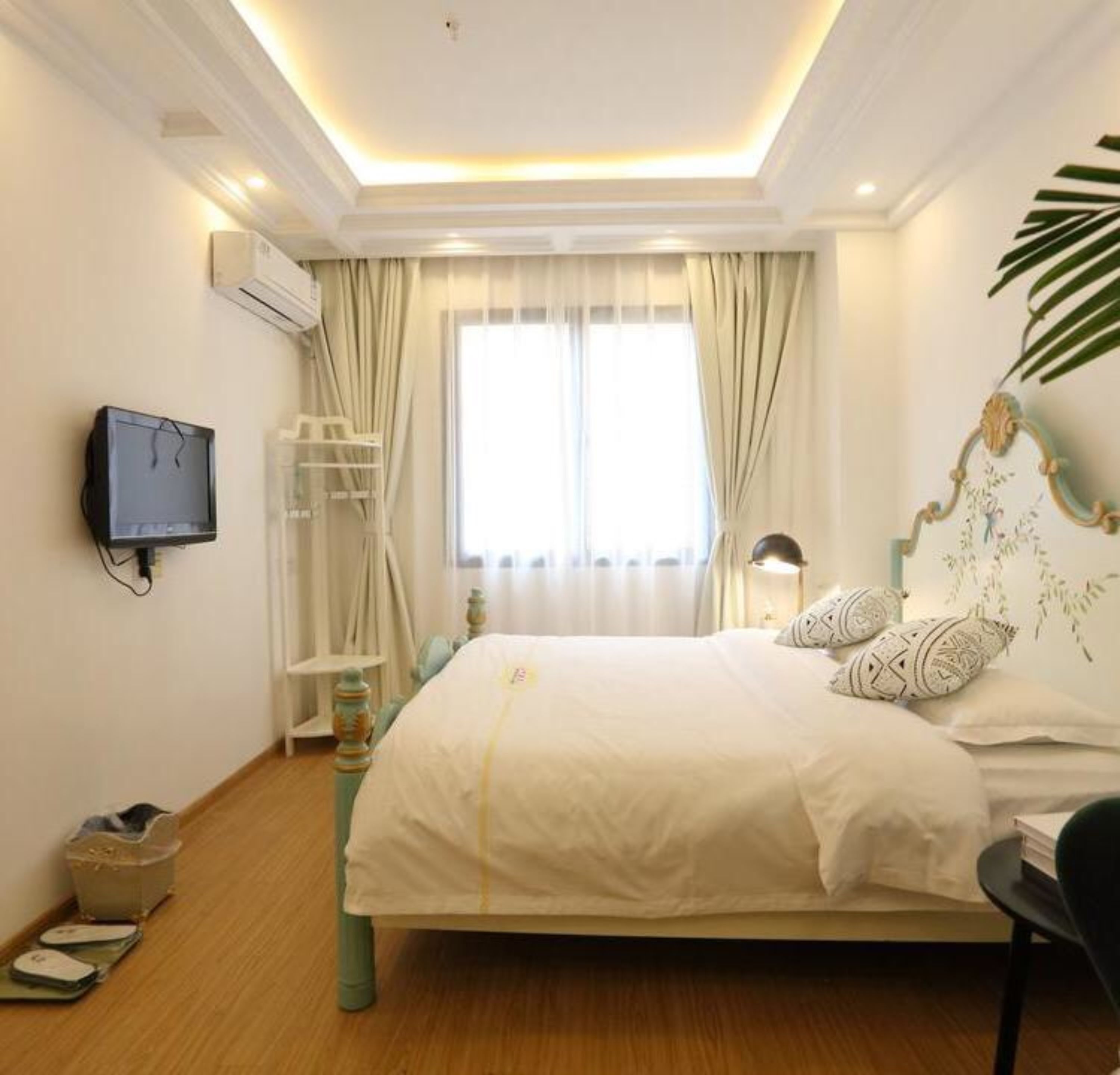 A hotel room with a bed, a TV mounted on the wall, a window with curtains, and a plant.