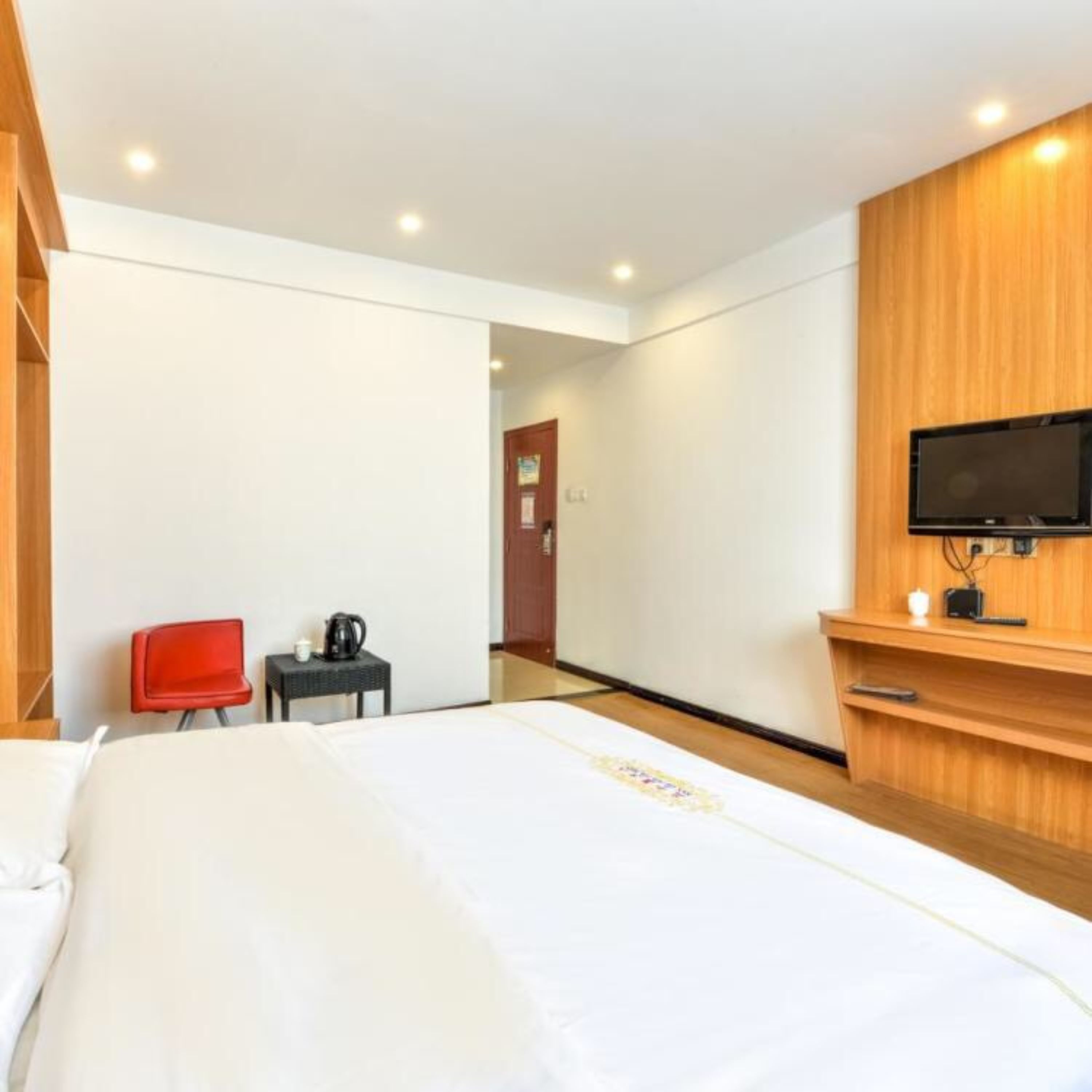 A hotel room with a large bed, a red chair, a small table with a kettle, and a TV mounted on a wooden panel.
