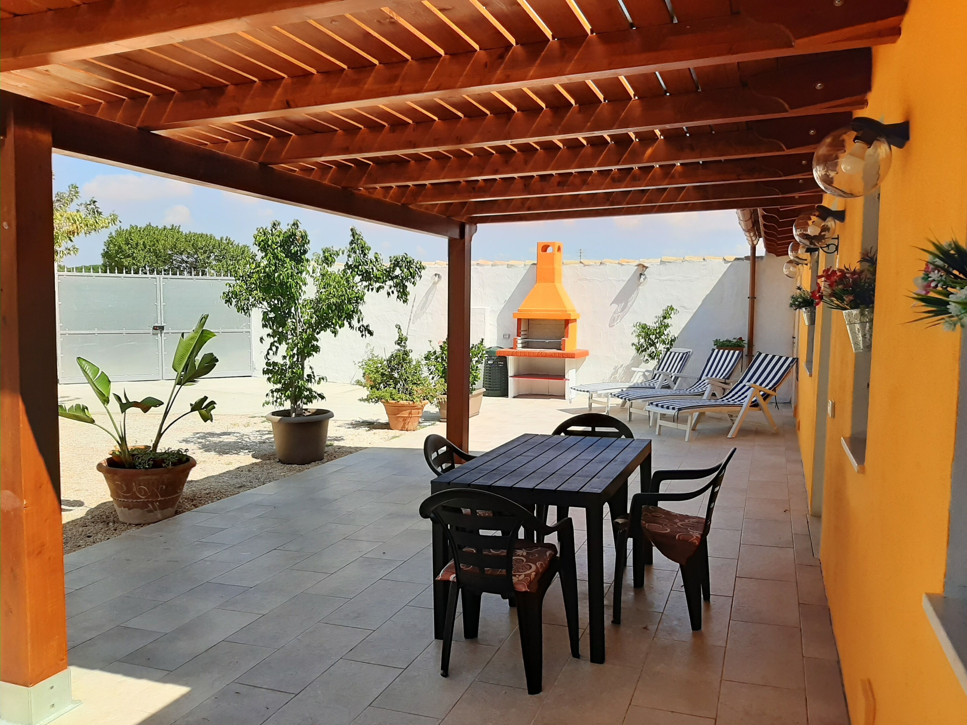 A covered patio with a wooden ceiling, a table with chairs, potted plants, and a barbecue grill.