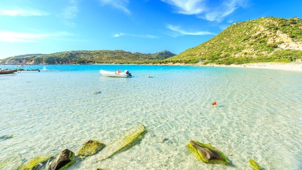Une plage dégagée avec des rochers au premier plan, un bateau et un paysage vallonné en arrière-plan.