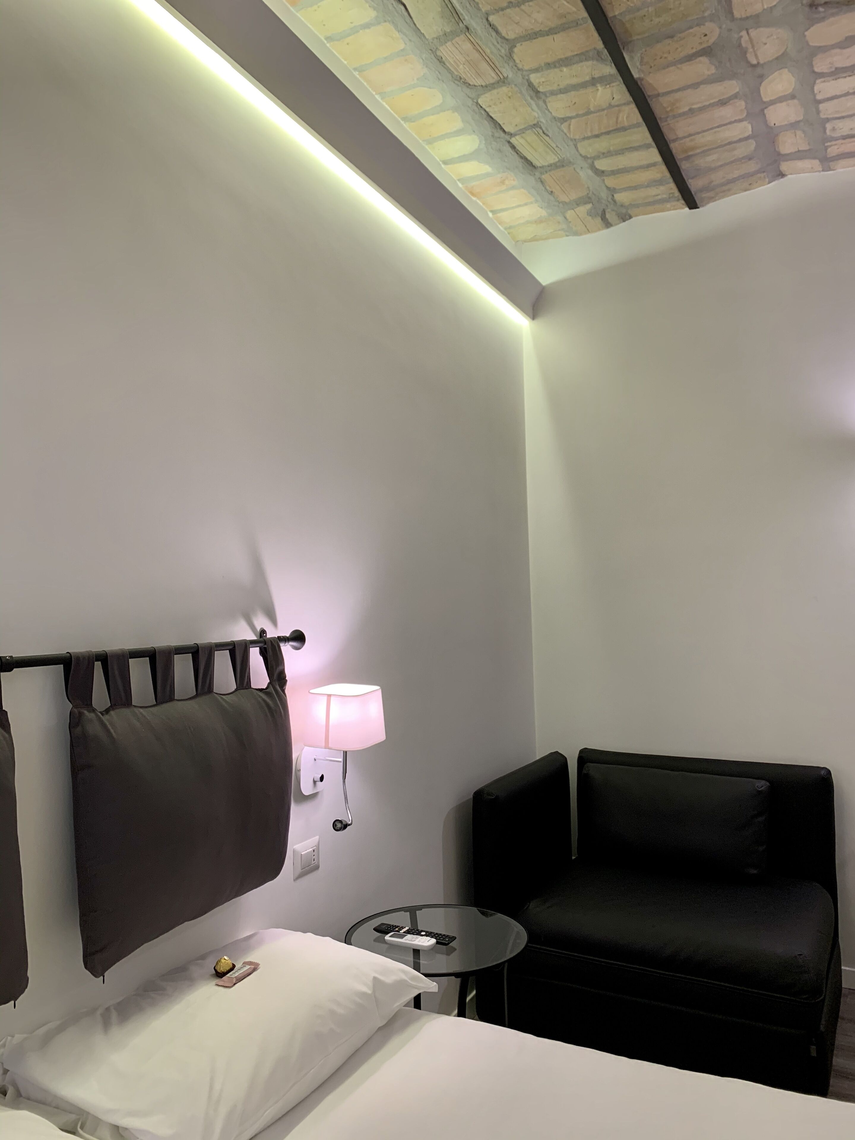 Une chambre d’hôtel avec un lit, une petite table ronde avec une lampe, un fauteuil noir et un plafond dont on peut voir la maçonnerie en briques apparentes.