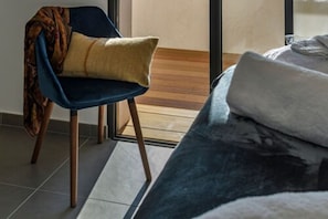 Una habitación moderna con una silla azul oscuro, un cojín color beige y una cama grande con una manta blanca.
