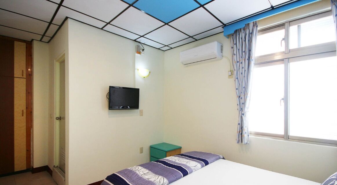 A bedroom with a bed, a television, an air conditioning unit, and a window with curtains.