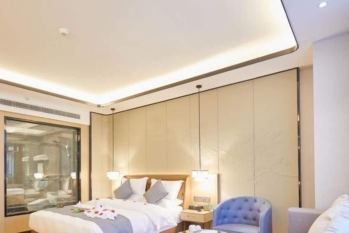 A modern hotel room with a large bed, a seating area with a coffee table, and a large window with blinds.