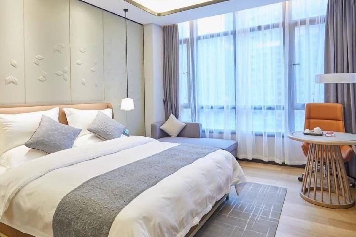 A modern hotel room with a large bed, a small round table, and a view of the city through large windows.