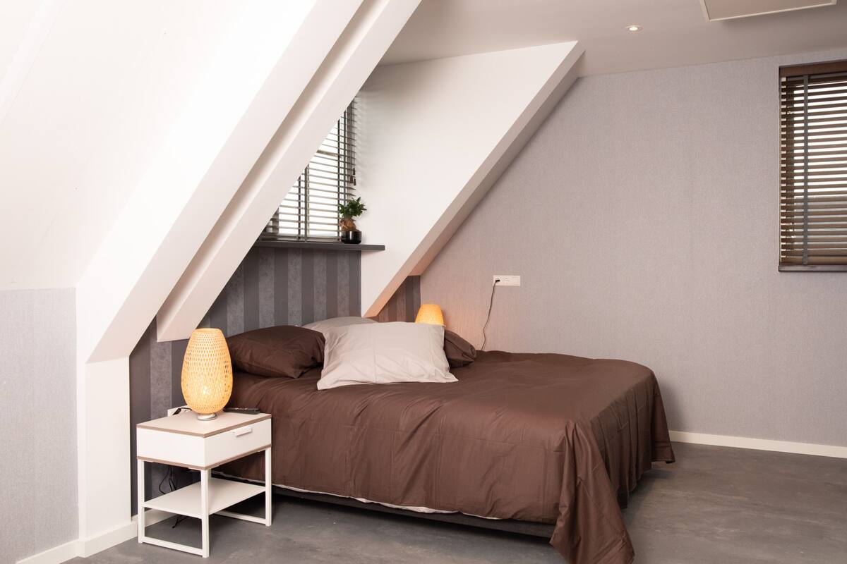 A modern bedroom with a sloped ceiling, a bed with brown bedding, a bedside table with a lamp, and a small plant on the nightstand.