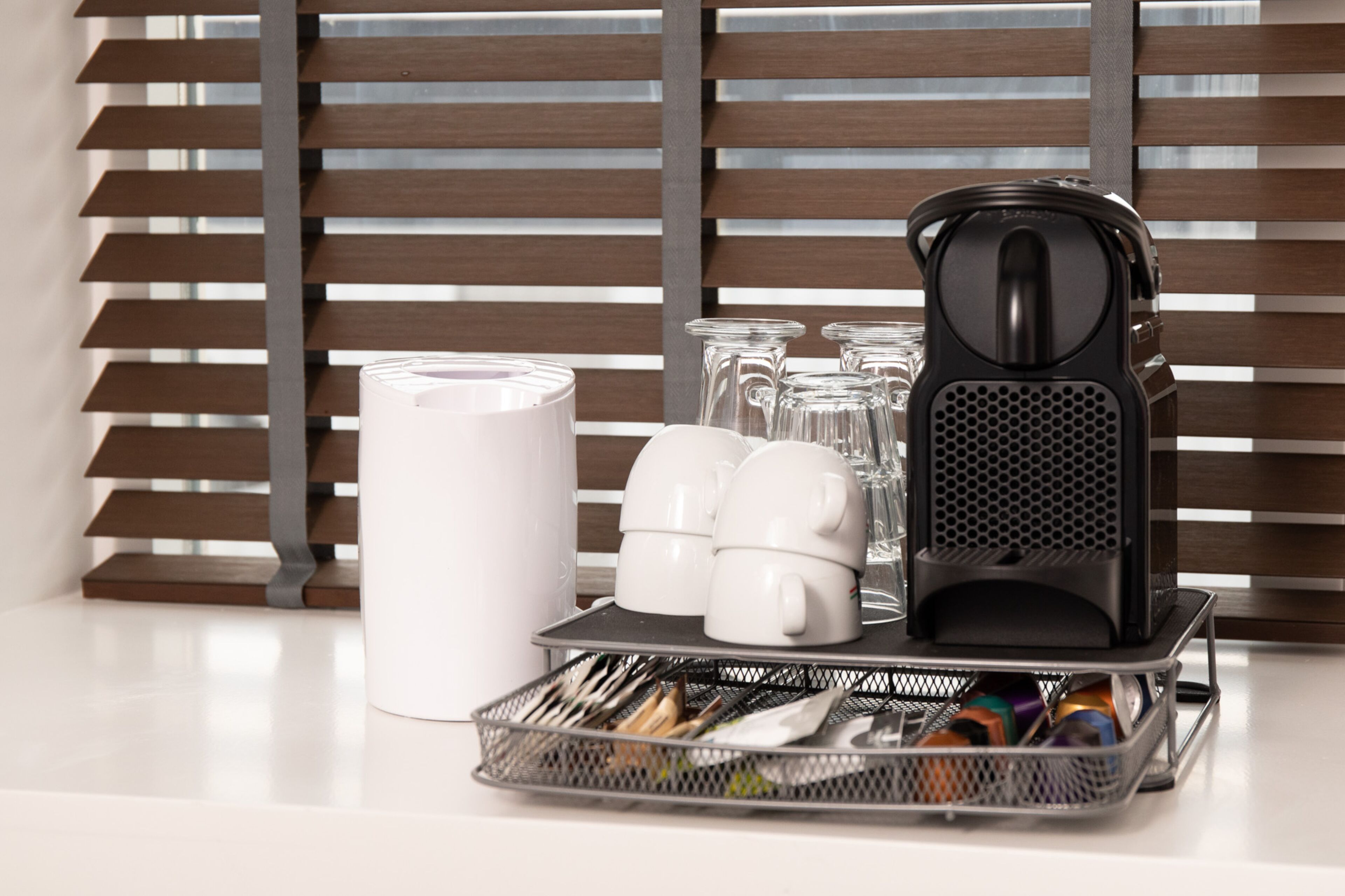 Un plan de travail de cuisine avec une machine à café, une tasse blanche, une carafe en verre et un panier rempli d’ustensiles.