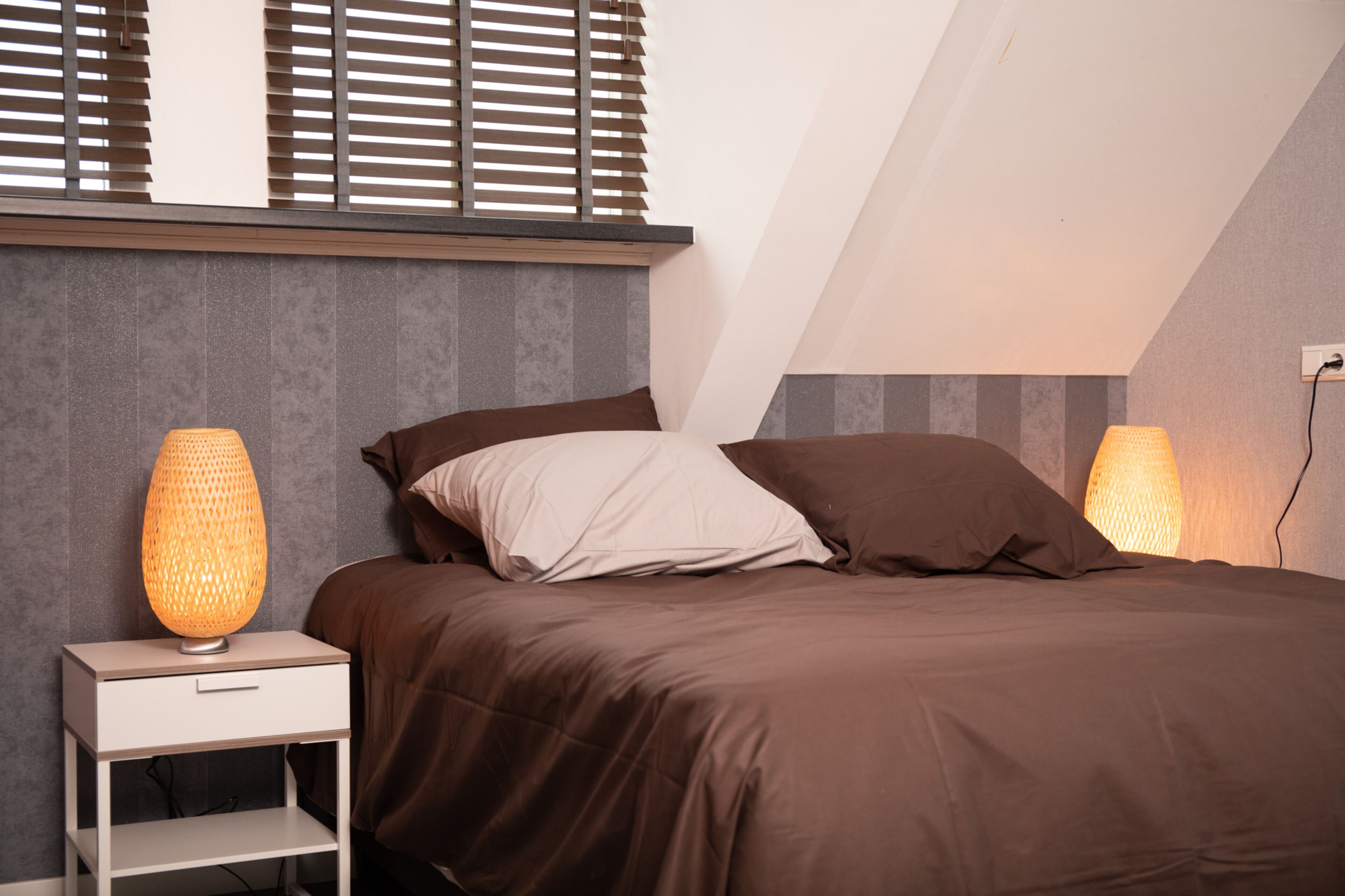 A bedroom with a bed, two bedside lamps, a nightstand, and a window with blinds.