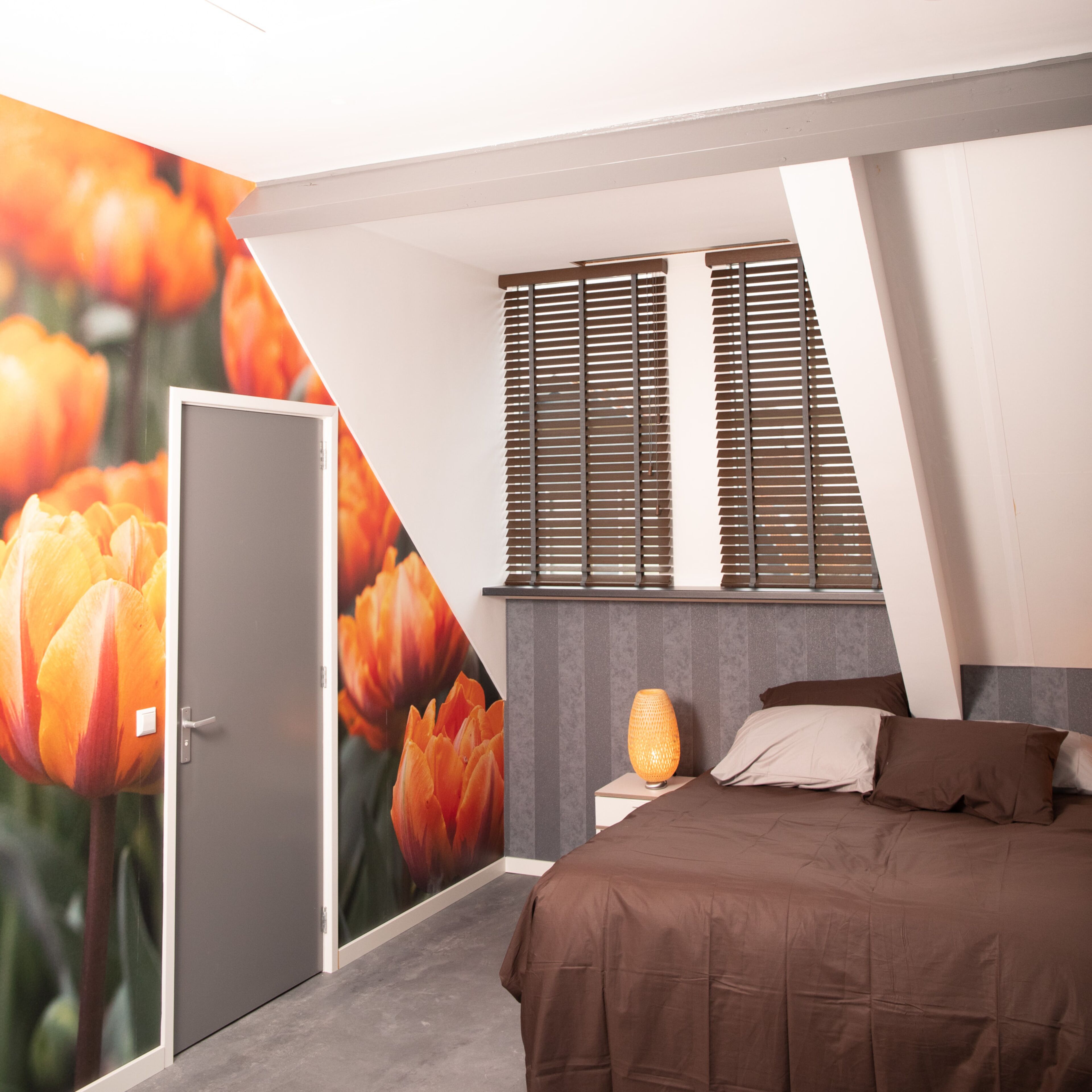 A bedroom with a bed, a nightstand, a lamp, a window with blinds, and a door.