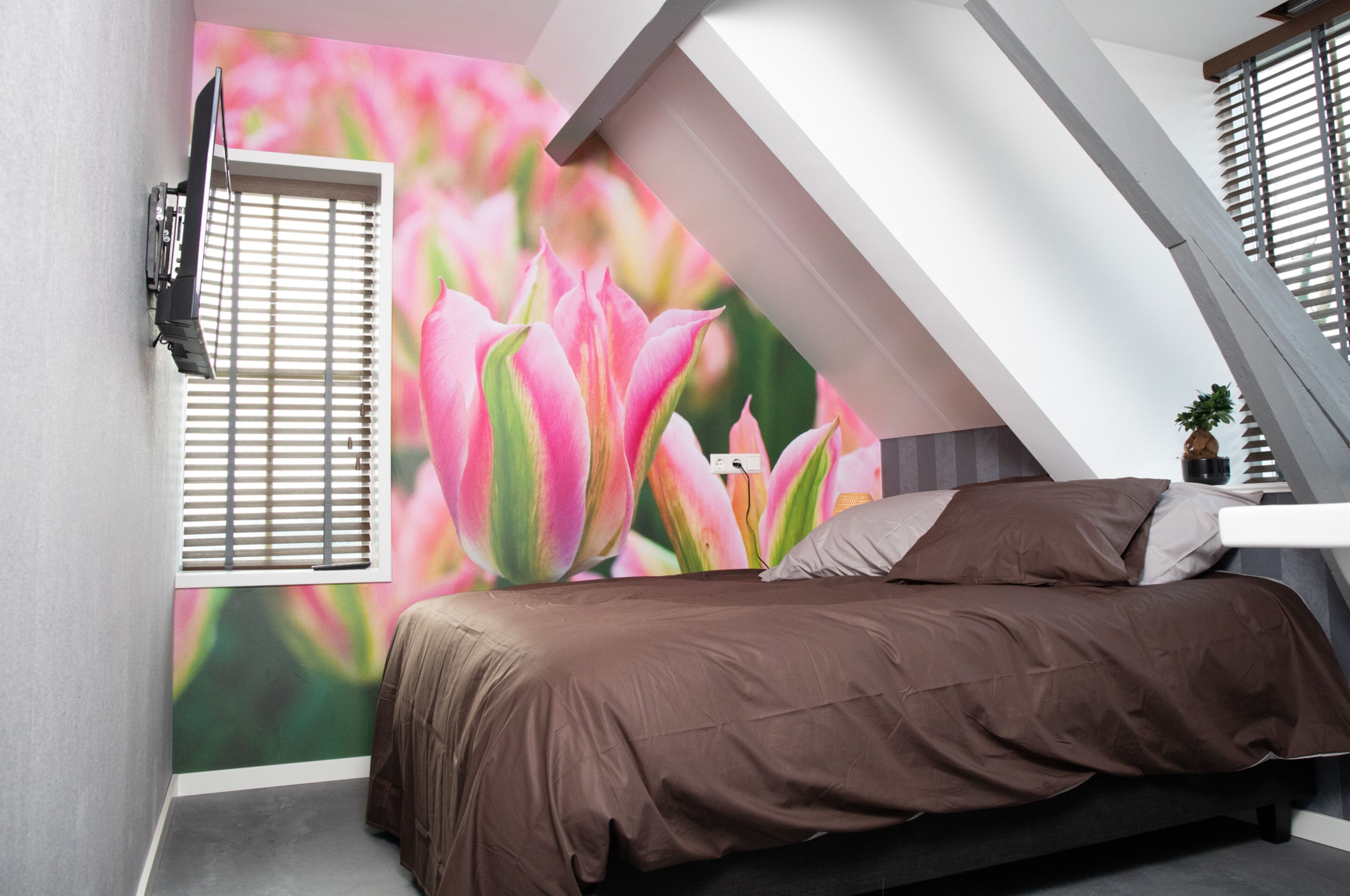 A bedroom with a sloped ceiling, a bed with brown bedding, a wall-mounted TV, and a window with blinds.