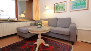 Ein Wohnzimmer mit grauem Sofa, rundem Couchtisch mit Pflanze, gemustertem Teppich und zwei gerahmten Bildern an der Wand.