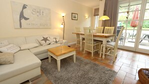 A living room with a sofa, coffee table, and dining area with chairs. There is a painting on the wall and a window with curtains.