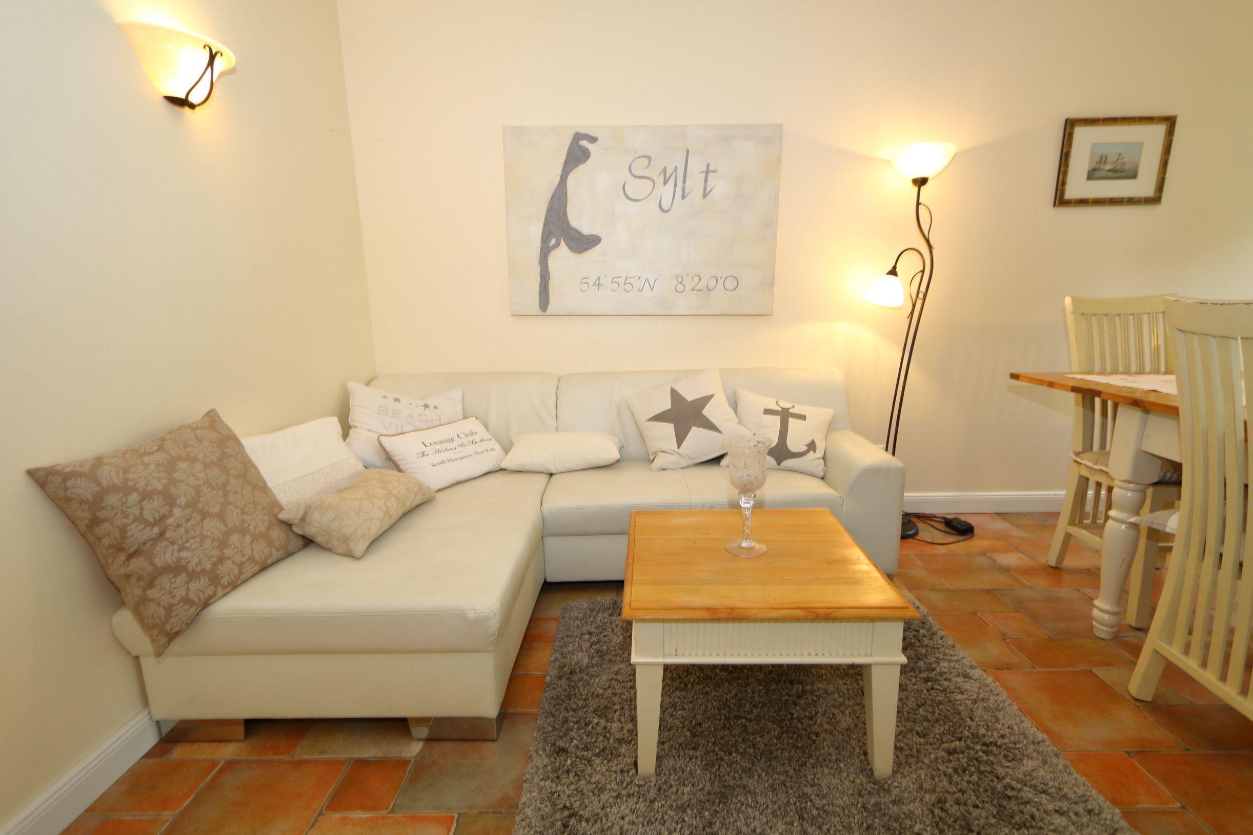 A living room with a white sofa, a wooden coffee table, and a rug. There is a wall-mounted artwork with the text "Slyt 5455'N 8200".