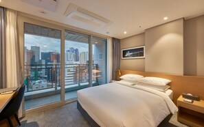 Ein Hotelzimmer mit Bett, Schreibtisch, Stuhl, Nachttisch und Blick auf die Stadt durch große Fenster.