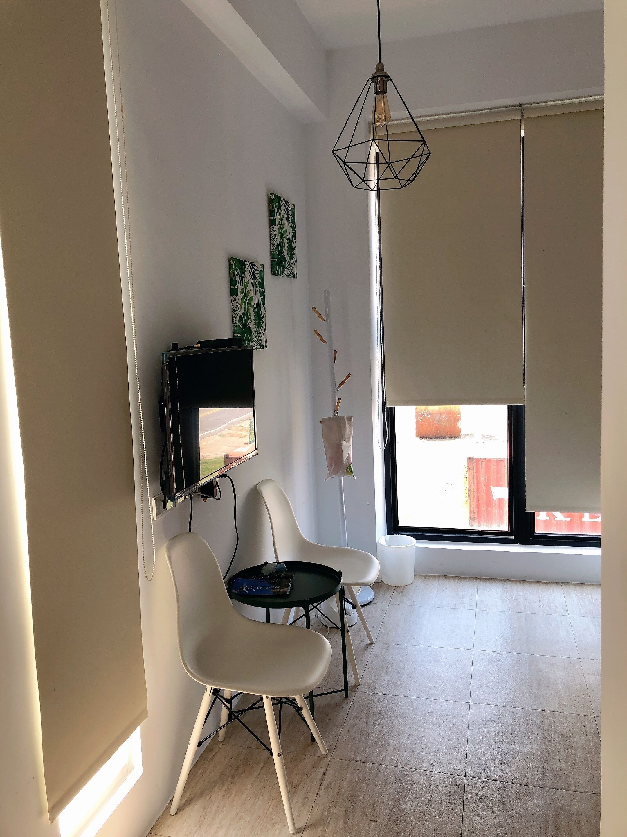 A modern living room with a flat-screen TV mounted on the wall, two white chairs, a small round table, a hanging light fixture, and a window with a view of the outside.